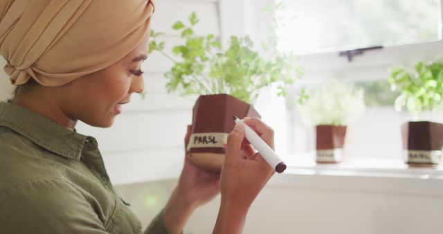 Image of biracial woman in hijab writing on herb pots. Lifestyle, houseplants, spending free time at home concept.