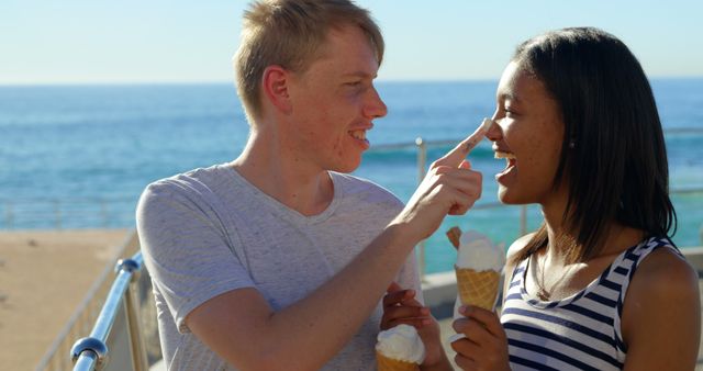 A young Caucasian man playfully touches the nose of a young African American woman with ice cream, with copy space. They share a joyful moment by the sea, highlighting a carefree and romantic connection.