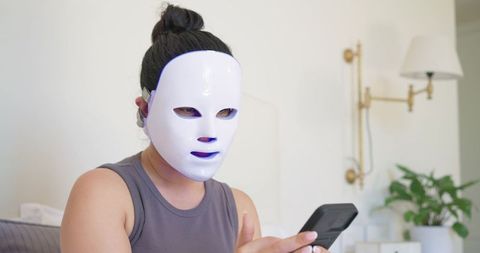 Woman wearing led facial mask while using smartphone on bed practicing at-home skincare routine