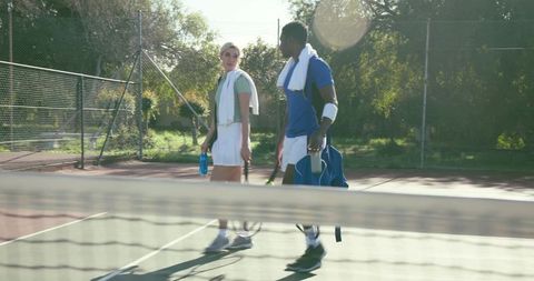 Tennis partners walking on sunlit outdoor court carrying rackets and towels after practice