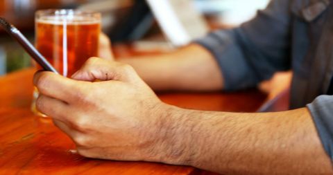 Scene of someone focusing on a smartphone with a beverage nearby captures a moment of leisure and communication. Perfect for use in articles about social media, modern lifestyle, technology, or advertising products such as mobile apps or beverages. The focus on hands emphasizes human interaction with technology in a relaxed, everyday environment.
