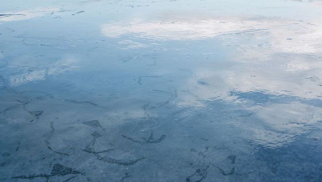 Thin ice sheet reflecting cloudy sky with cracks and meltwater ripples