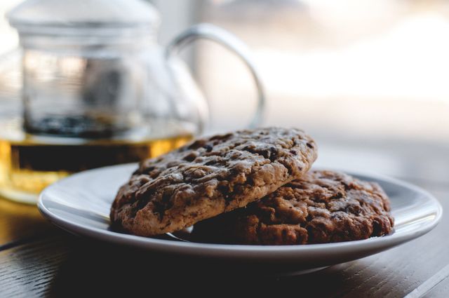 Perfect for advertising bakeries or coffee shops, this image highlights freshly baked oatmeal cookies placed on a white plate with a glass teapot in the blurred background. It conveys a homely and cozy atmosphere, ideal for promoting cozy environments, comfort foods, recipe blogs, culinary uses, kitchen decor, and home baking endeavors.