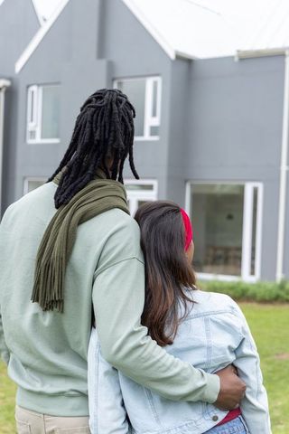 Diverse couple standing on front lawn embracing while looking at modern gray home
