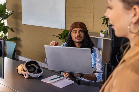 Coworkers discussing project while colleague working on laptop with vr headset in modern workspace