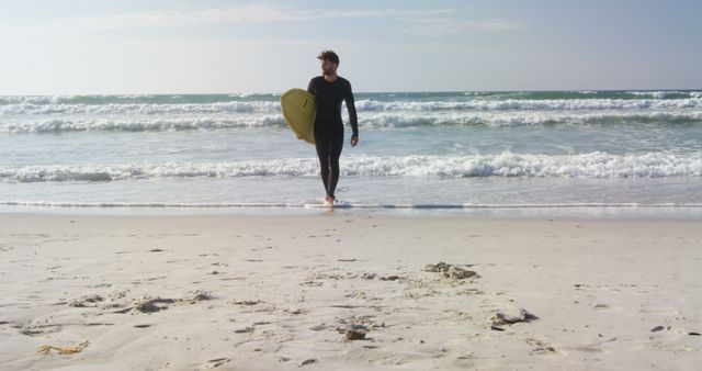 Male surfer walking on sandy beach while carrying surfboard. Sun is shining, waves crashing. Ideal for beach activities promotions, surfing lifestyle content, fitness and outdoor adventure themes.