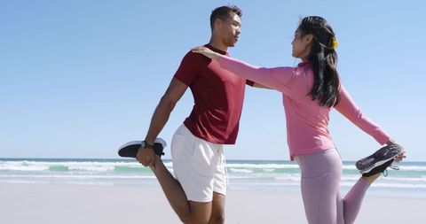 Couple Performing Partner Stretch on Sunny Beach Holding Sneakers While Balancing