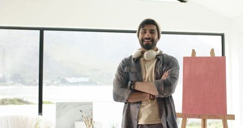 Confident indian artist standing in bright studio with easel and headphones by window