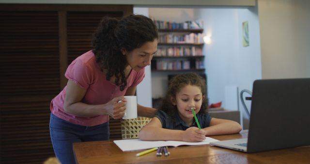 Mother supervising daughter doing homework at home. Young girl focuses on writing in notebook while using a laptop. Mother offers support, holding coffee mug, standing beside. Suitable for education, family, parenting, remote learning content.