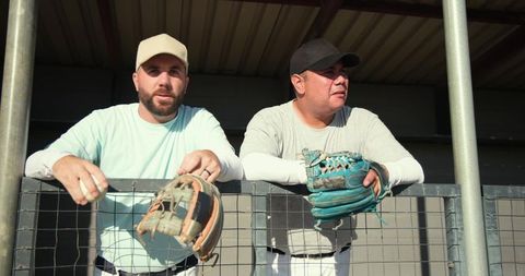 Diverse teammates leaning on dugout fence holding gloves in sunny ballpark