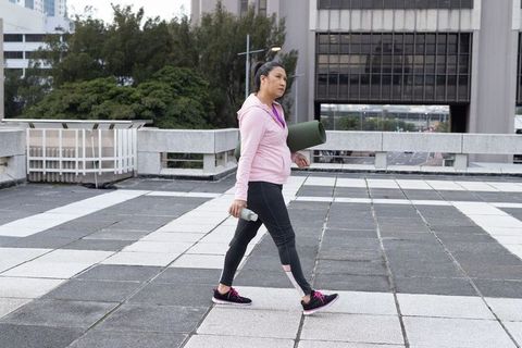 Mature woman walking with yoga mat and water bottle across urban rooftop plaza for fitness