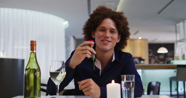 Biracial man having romantic dinner at restaurant holding red rose and talking to camera. romantic evening out celebrating valentine's day.