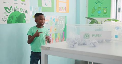 Young african american boy holding plastic bottle pointing at paper recycling bin in eco class