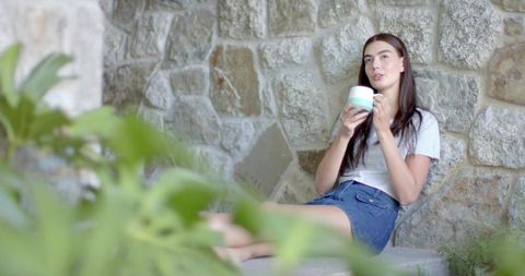Young woman relaxing with pastel mug on stone bench in sunlit courtyard greenery