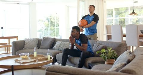 Intense game-watching friends holding basketball and sharing pizza in sunlit modern living room