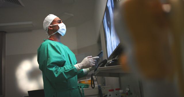 Healthcare professional wearing surgical cap, mask, and gloves examining medical scans on screen in hospital setting. Useful for illustrating medical diagnosis, patient care, hospital environments, healthcare technology, and professional healthcare workers in action.