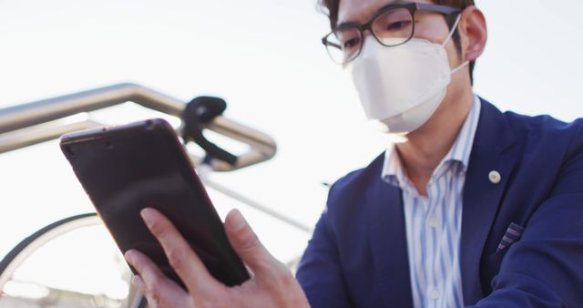 Asian man wearing face mask using digital tablet while sitting on the stairs at corporate park. health protection and safety during covid-19 pandemic concept
