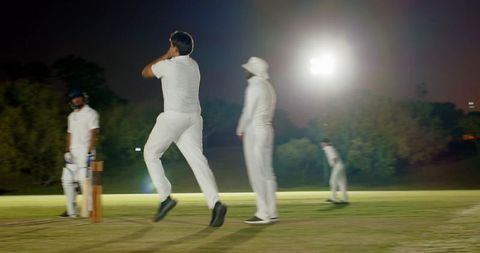 Night cricket under floodlights with bowler leaping, batsman at crease, diverse team