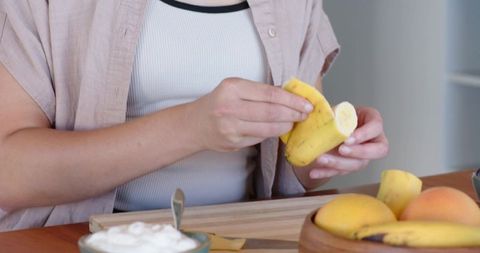 Peeling banana at cozy breakfast table holding fruit over cutting board with yogurt bowl