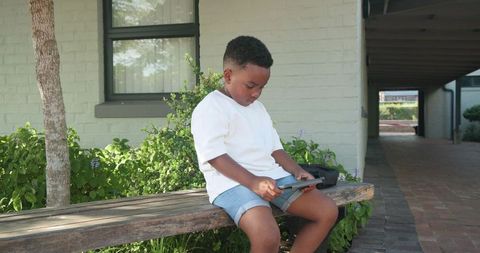 African american boy sitting on wooden bench using tablet outdoors on porch with backpack