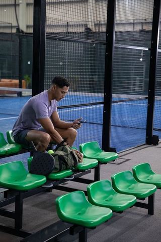 Man checking phone courtside at indoor padel arena with green seating