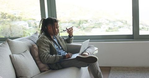 African american man working at home on laptop while holding handheld device by window