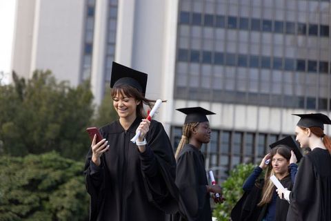 Female college graduates celebrating outdoors holding diplomas and checking smartphone
