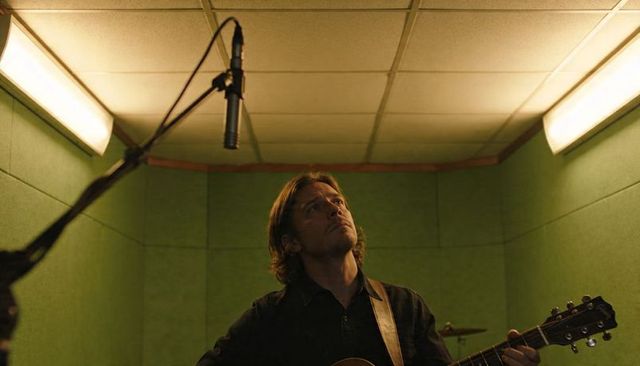 Contemplative singer-songwriter recording acoustic guitar in green vocal booth near mic