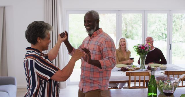 Senior couple enjoying spontaneous dance in living room with friends smiling at dining table. Ideal for content about happy aging, friendship, family gatherings, and celebrating life. Great for lifestyle, social interactions, and elderly wellness themes.