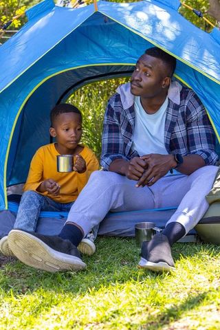 African american father and son camping together at tent entrance sipping from metal mugs