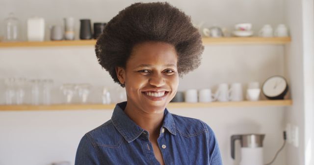 An image depicts a cheerful woman with natural afro hair, dressed in a casual denim shirt, standing in a kitchen. Shelving with glassware and a minimalist decor is seen in the background. Ideal for use in blogs about home living, lifestyle magazines, advertisements promoting casual or home settings, and content focusing on positive portrayals of natural beauty and confidence.