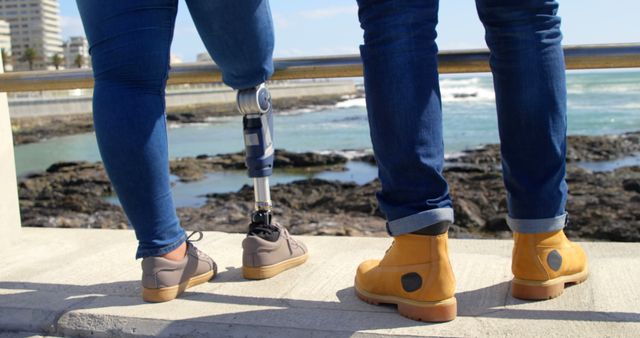 Two friends standing by coastal rail, one with prosthetic leg. Ideal for themes of friendship, inclusion, disability awareness, outdoor activities.