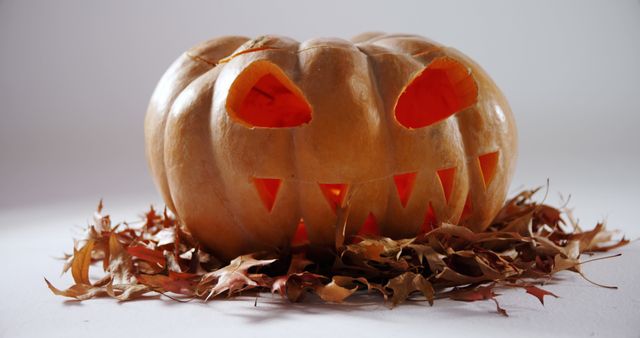 A carved pumpkin with a spooky face sits amidst dry autumn leaves, symbolizing Halloween festivities. Its glowing eyes and jagged smile create an eerie atmosphere for the holiday celebration.