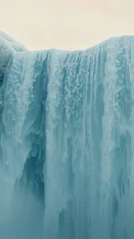 Vertical video showing clouds casting light across frozen waterfall cliff with rising mist
