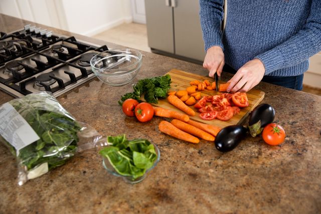 Woman chopping fresh vegetables including carrots, tomatoes, and eggplants on a wooden board in a modern kitchen. Ideal for use in articles or advertisements about home cooking, healthy eating, meal preparation, and lifestyle changes during quarantine and social distancing.