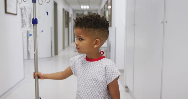 A young male patient holding an IV stand while standing in a sterile hospital corridor. Suitable for use in healthcare promotions, pediatric care advertisements, health insurance marketing, and raising awareness about children's health issues.