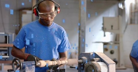 Black craftsman shaping wooden spindle on lathe in industrial workshop wearing safety gear