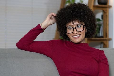 Smiling woman with curly afro and black glasses relaxing on gray sofa in cozy living room
