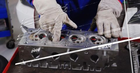 Technician inspecting cylinder head on workbench, gloved hands pointing at valve seats for rebuild