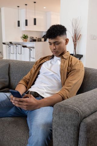 Young man scrolling smartphone on couch in modern open-plan apartment with minimalist interior