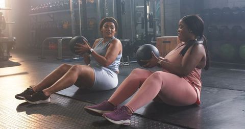 Diverse women performing partner core workout with medicine balls in gym
