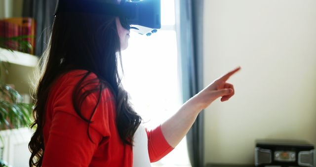 This image shows a young adult using a virtual reality headset in a brightly lit living room. They are pointing, likely interacting with a VR environment. It is suitable for themes related to VR technology, home entertainment, immersive experiences, and modern technology adoption.