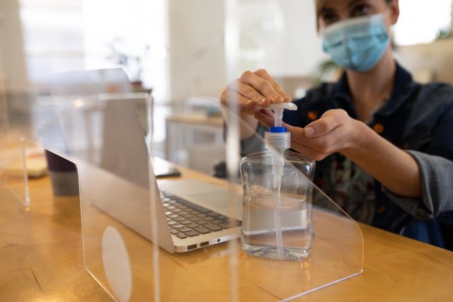 Caucasian woman wearing a facemask pumping sanitizer onto her hands at her desk in front of her laptop. desks in the office are separated with a transparent plastic divider.