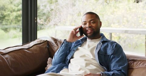 African american man relaxing on leather sofa talking on smartphone by window in daylight