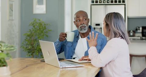 Senior Multiracial Couple Discussing Finances at Kitchen Table with Laptop and Coffee