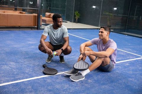 Diverse male friends resting on indoor blue padel court in sportswear holding paddles