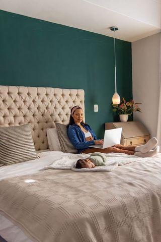 Working from bed while baby sleeping on cozy neutral bedding, mother using laptop