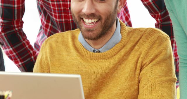 Image features a young man smiling while working on his laptop, surrounded by colleagues in a modern office setting. Ideal for use in business, coworking, teamwork, and technology-related presentations, websites, and promotional materials reflecting a casual, collaborative work environment.