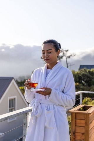 Asian woman in white robe enjoying morning tea on misty balcony overlooking hills