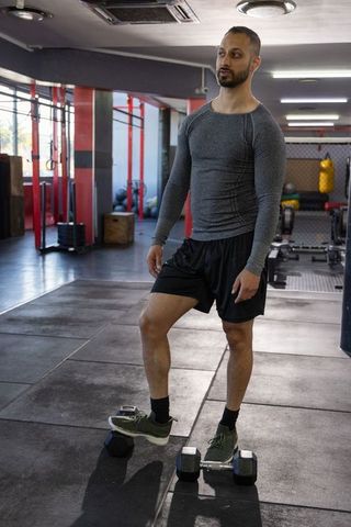 Indian man standing on dumbbells in gym wearing gray long sleeve top and athletic shorts